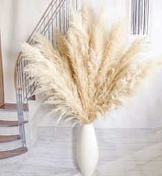 a white vase filled with dry grass on top of a wooden floor next to a stair case