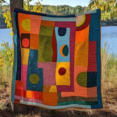 a multicolored quilt hanging from a tree by the water with trees in the background