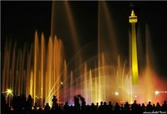 people are standing in front of a fountain at night with lights and water spraying from it