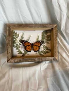 an orange butterfly sitting in a wooden frame on a white cloth covered bedding area