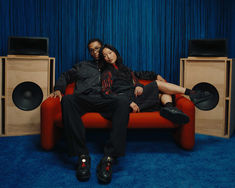 a man and woman sitting on a red couch in front of two speaker system's