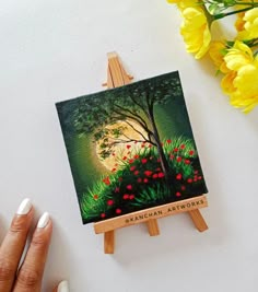 a painting on an easel being held by a woman's hand with yellow flowers in the background