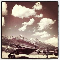 an old airplane sitting on the ground with mountains in the background and clouds in the sky