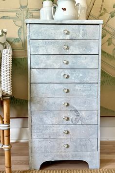 an old white dresser with drawers in front of a wallpapered wall and vase