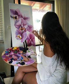 a woman sitting in front of a painting on a easel with flowers painted on it