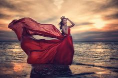 a woman in a red dress is standing on the beach with her arms behind her head