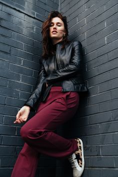 a woman leaning against a brick wall with her hands on her hips, wearing red pants and a black leather jacket