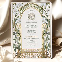 an ornate wedding card with white flowers and gold trimmings on the front, sitting next to some feathers