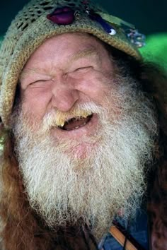 an old man with long hair and a beard wearing a knitted hat smiles at the camera