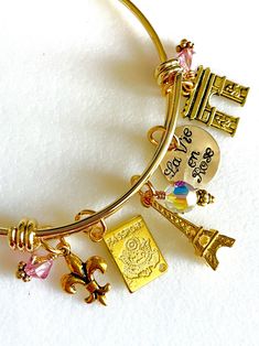 "Perfect souvenir of an enchanting trip to Paris, France. Adjustable gold plated bangle is highlighted with hand stamped quote \"La Vie en Rose\" and 5 detailed Parisian charms accented with rose colored Swarovski crystals AB.  Adjustable bangle is made of heavy gold plate over 1.8mm stainless steel. It is hypoallergenic and tarnish resistant. The diameter is 2 1/2\" to fit most.  Hand stamped 5/8\" 24 gauge shiny brass disc reads \"La Vie en Rose\". Two 6mm rose colored Swarovski crystals AB an Personalized Pink Bangle Charm Bracelet, Nickel-free Pink Bangle Charm Bracelet, Nickel Free Pink Bangle Charm Bracelet, Honeymoon Paris, Fun Halloween Earrings, 3d Eiffel Tower, Paris Charm Bracelet, Paris Bracelet, Paris Souvenirs