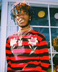 a young man with dreadlocks standing in front of a window wearing a red and black striped shirt