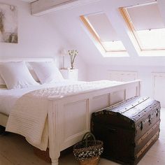 a white bed sitting under two windows next to a wooden trunk and basket on the floor