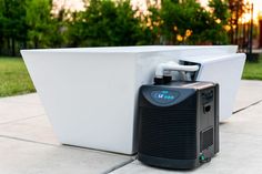 an air conditioner sitting on the ground next to a white wall and green trees