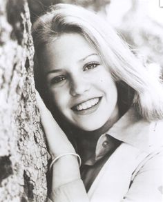 a black and white photo of a smiling woman leaning against a tree with her hand on her head