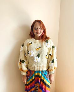 a woman standing in front of a wall wearing a sweater and colorful skirt with flowers on it