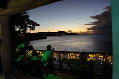 some people are sitting at a table by the water with lights on it and there is a sunset in the background