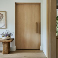 a wooden door in a white room next to a small table with flowers on it