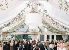 a group of people standing under a white tent at a formal event with tables and chairs