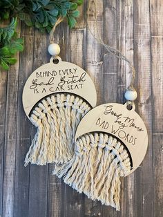 two tasseled ornaments with words on them hanging from a wooden wall next to greenery