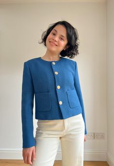 a woman standing in front of a white wall wearing a blue jacket and cream pants