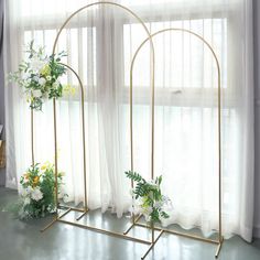 two gold arch stands with flowers and greenery on them in front of a window