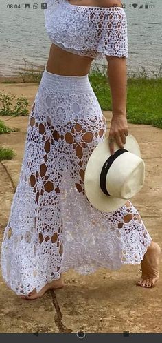 a woman in a white crochet dress and hat walking on the sidewalk near water