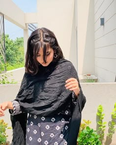 a woman wearing a black shawl standing in front of a building with plants and bushes