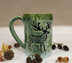 a green mug with a deer on it surrounded by pine cones and other autumn decorations