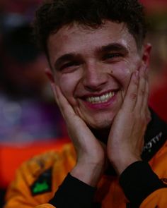 a smiling young man with his hands on his face, looking at the camera while wearing an orange and black jacket