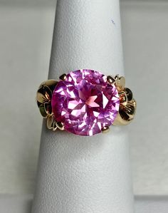 This ring is beautiful! The ring features a nice quality synthetic pink spinel that is 11mm in diameter. The ring is in nice condition for its age, but will show signs of natural wear (see photos). The ring is solid 10k yellow gold and weighs a hefty 4.5g Fine Jewelry Pink Ruby Ring With Round Band, Pink Ruby Ring With Round Band In Fine Jewelry, Pink Ruby Ring With Diamond Accents, Pink Topaz Promise Ring In Round Shape, Pink Round Topaz Promise Ring, Pink Topaz Promise Ring With Round Shape, Pink 14k Gold Birthstone Ring, Pink Round Ruby Promise Ring, Pink 14k Stamped Promise Ring