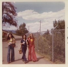 three women standing on the side of a road next to a chain link fence with trees in the background