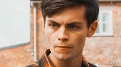 a close up of a person wearing a plaid shirt and looking at the camera with a brick building in the background