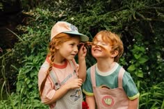 two young children standing next to each other in front of some bushes and trees with their eyes closed