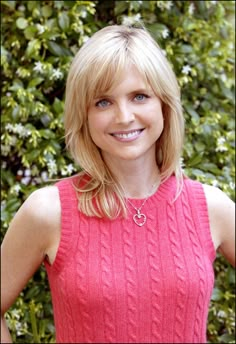 a woman with blonde hair wearing a pink sweater and smiling at the camera while standing in front of some bushes