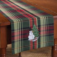 a plaid table runner with a snowman and christmas tree embroidered on the front, sitting on a wooden dining room table