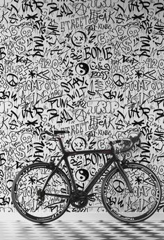 a black and white photo of a bike in front of a wall with graffiti written all over it