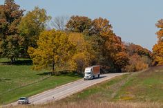 two trucks are driving down the road in front of trees with yellow and orange leaves