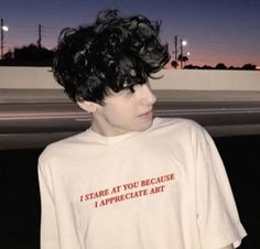 a young man with black hair wearing a white t - shirt that says i stare at you because i appreciate art