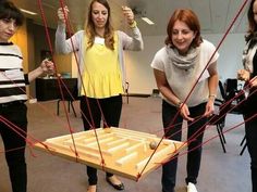 three women standing in front of a table made out of sticks and wires, with one woman holding up a piece of wood