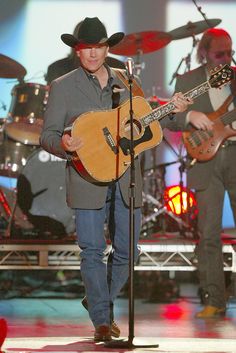 a man standing in front of a microphone while holding a guitar and wearing a cowboy hat