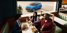 three people sitting at a table with food and drinks in front of a blue car