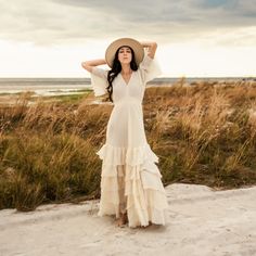 a woman in a white dress and hat standing on the beach with her arms behind her head