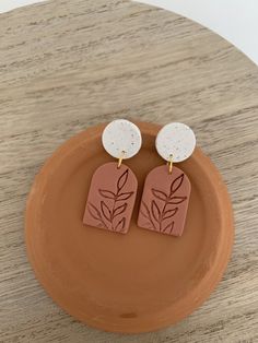 pair of earrings with white disc and brown leaf design on wooden plate sitting on top of wood table