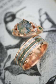 A matching wedding ring set from Staghead Designs sits on an antique bird illustration. First is our "Lucy In The Sky" design engagement ring in 14K rose gold with a hexagon cut moss agate center stone, diamond halo & accents, & moss inlays. Below is "Rio" in 14K rose gold with a centered crushed moldavite inlay & a damascus steel inlay on either side. These rings are fully customizable & can be made with a wide variety of unique materials & stones. Labrodite Engagement Ring, Make Wedding Rings, Moss Agate Matching Rings, Wedding Rings With Green Stones, Libra Engagement Ring, Matching Moss Agate Wedding Rings, Moldavite Engagement Ring, Womens Engagement Rings Unique, Unique Engagement Rings Color