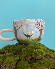 a coffee cup sitting on top of a moss covered ground with trees and dinosaurs painted on it