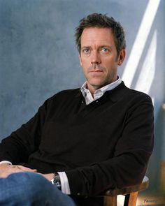 a man sitting in a chair with his arms crossed looking at the camera while wearing a black sweater and blue jeans