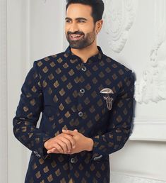 a man with a beard wearing a blue and gold patterned shirt standing in front of a white wall