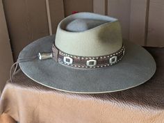 The Bandit is a stand out hat that captures the feeling of the American West. . The base of the hatband is distressed leather that is then layered with imprinted brown leather, bolo sliders and lots of nailspots. It is finished with this colorful feather combo. The brim of the LID custom hat body is 4" in diameter and the crown is 4.5". This is a stiff hat. Available in Putty or Pecan All hats are made to order. Allow up to 4 weeks for your hat to ship. Rugged Distressed Brown Leather Hat, Rustic Hat With Distressed Brown Curved Brim, Rugged Flat Brim Hat For Ranch, Rustic Hat With Curved Brim In Distressed Brown, Rustic Hat With Distressed Brown Color And Curved Brim, Rustic Distressed Brown Hat With Curved Brim, Rugged Flat Brim Ranch Hat, Distressed Brown Fedora For Country Events, Distressed Brown Leather Brimmed Hat