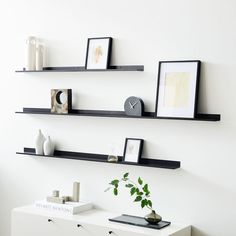 three black floating shelves on the wall above a white dresser with a plant and vase