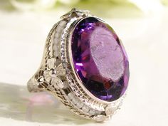 This sweet antique style engagement ring with a large amethyst colored glass stone has been well loved over the years and can still grace the finger of some vintage lover as an eye-catching statement ring! It is crafted in 14k white gold with orange blossom detailing and openwork filigree replicating the romantic Edwardian era. The large amethyst colored glass is oval cut measuring approximately 20mm long by 15mm wide or 14.96 carats. The dark purple colored glass stone with red flashes is bezel Antique Style Engagement Rings, Pearl Wedding Ring, Filigree Ring Gold, Timeless Engagement Ring, Floral Filigree, Traditional Engagement Rings, Antique Wedding Rings, Beautiful Wedding Rings, Vintage Engagement Ring